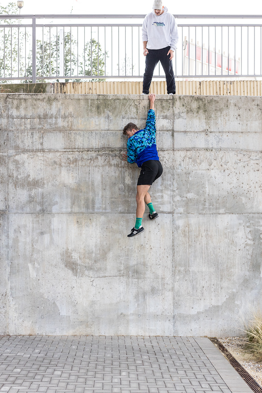 Zwei Personen trainieren Parkour im Outdoor-Bereich – eine schaut zu, die andere klettert an einer Wand hoch.